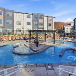 Outdoor apartment pool area with lounge chairs, a shaded seating structure, and modern beige-gray buildings nearby.