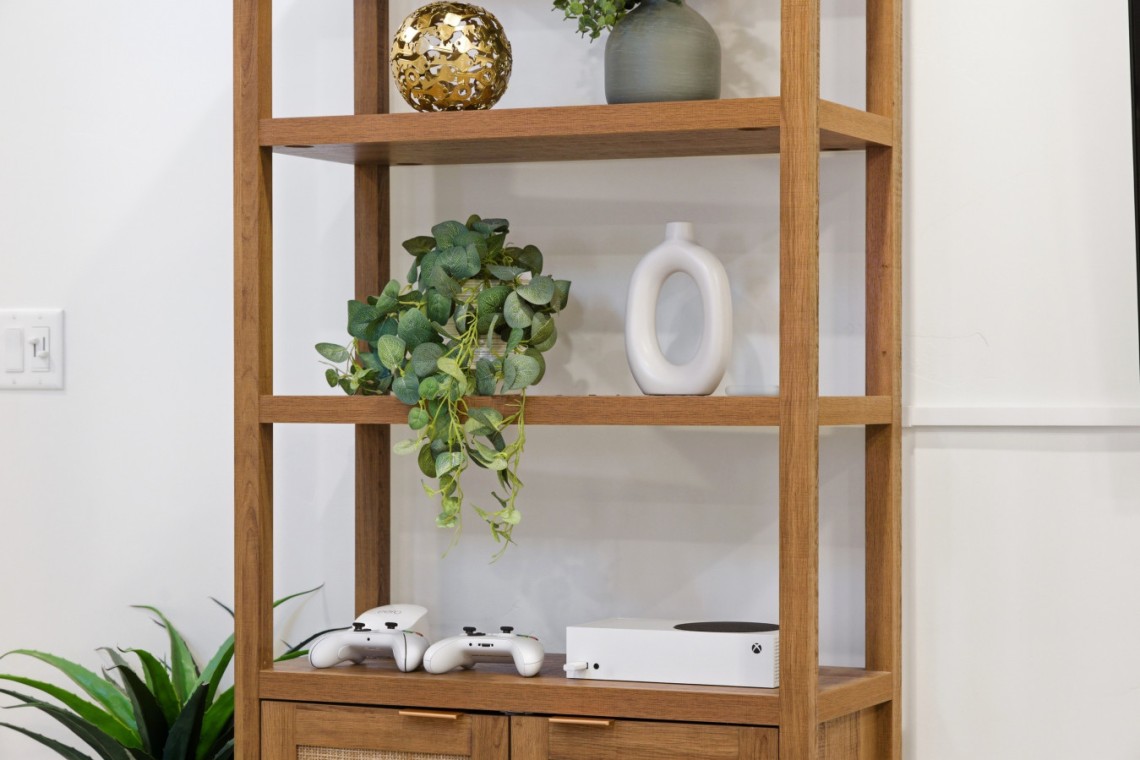 Wooden open-shelving unit with a trailing green plant, a white oval vase, and an Xbox console with controllers on the lower shelf.