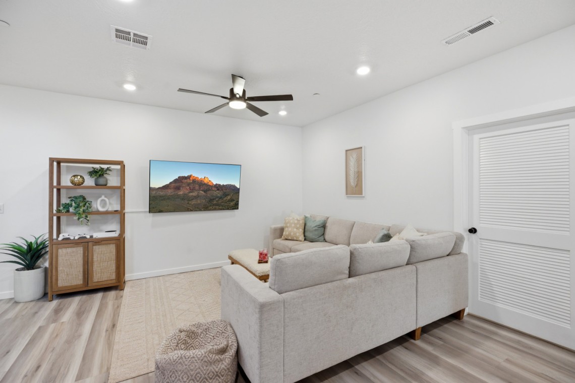 Living room with a light gray sectional sofa, wall-mounted TV, and a wooden shelf against a white wall.