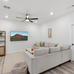 Living room with a light gray sectional sofa, wall-mounted TV, and a wooden shelf against a white wall.