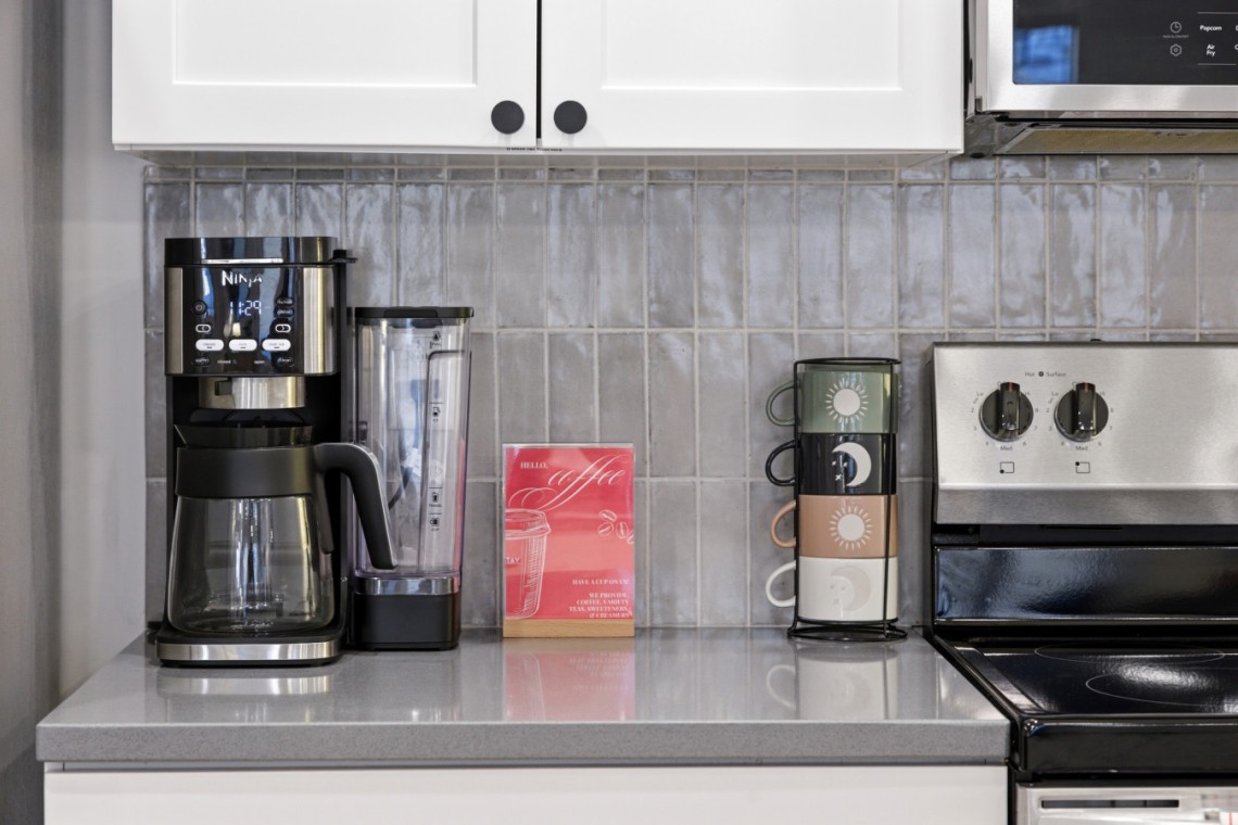 Countertop kitchen scene with a black drip coffee maker, a blender, a red 'Hello Coffee' card, and a stack of four mugs beside a stainless stove.