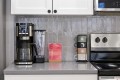 Countertop kitchen scene with a black drip coffee maker, a blender, a red 'Hello Coffee' card, and a stack of four mugs beside a stainless stove.