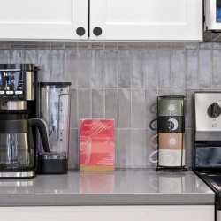 Countertop kitchen scene with a black drip coffee maker, a blender, a red 'Hello Coffee' card, and a stack of four mugs beside a stainless stove.