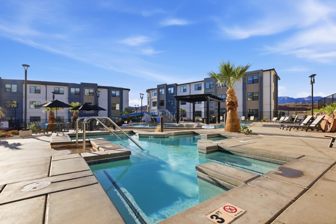 Outdoor pool area at a modern apartment complex with lounge chairs, palm trees, and blue sky.