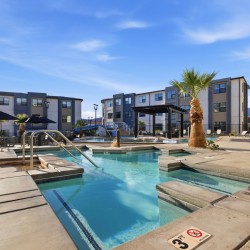 Outdoor pool area at a modern apartment complex with lounge chairs, palm trees, and blue sky.