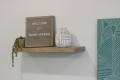 Wooden wall shelf with a sign reading 'WELCOME TO SUNNY SPRINGS', a white geometric vase, and a trailing succulent in a yellow pot.