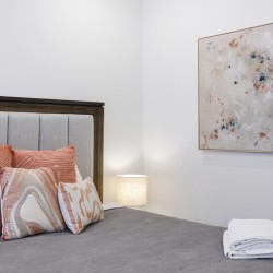 Bedroom scene with a gray upholstered bed, rust-orange and patterned pillows, and a folded white towel on a gray coverlet.