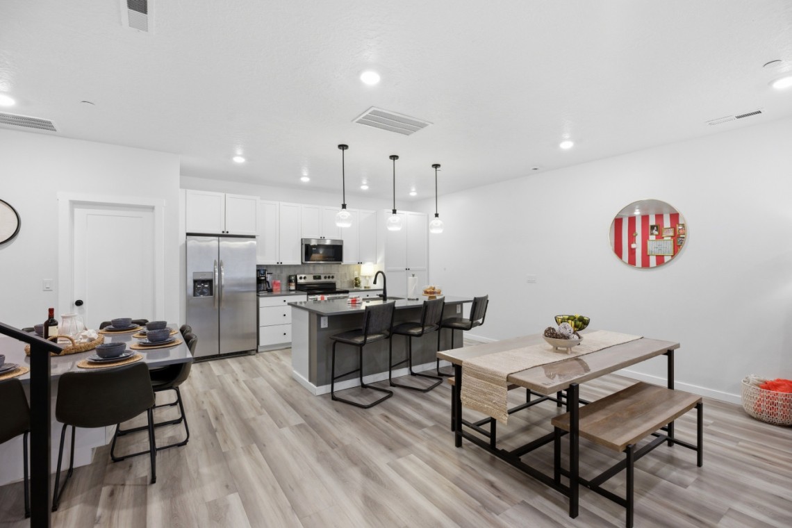 Open-concept kitchen and dining area with white cabinets, stainless fridge, island with black stools, and a wooden dining table on the right.