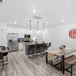 Open-concept kitchen and dining area with white cabinets, stainless fridge, island with black stools, and a wooden dining table on the right.