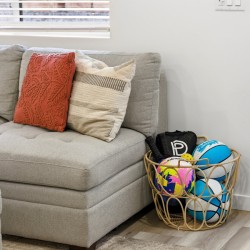 Grey fabric sofa with an orange patterned throw pillow and a beige striped pillow, next to a wicker basket filled with colorful sports balls by the door.