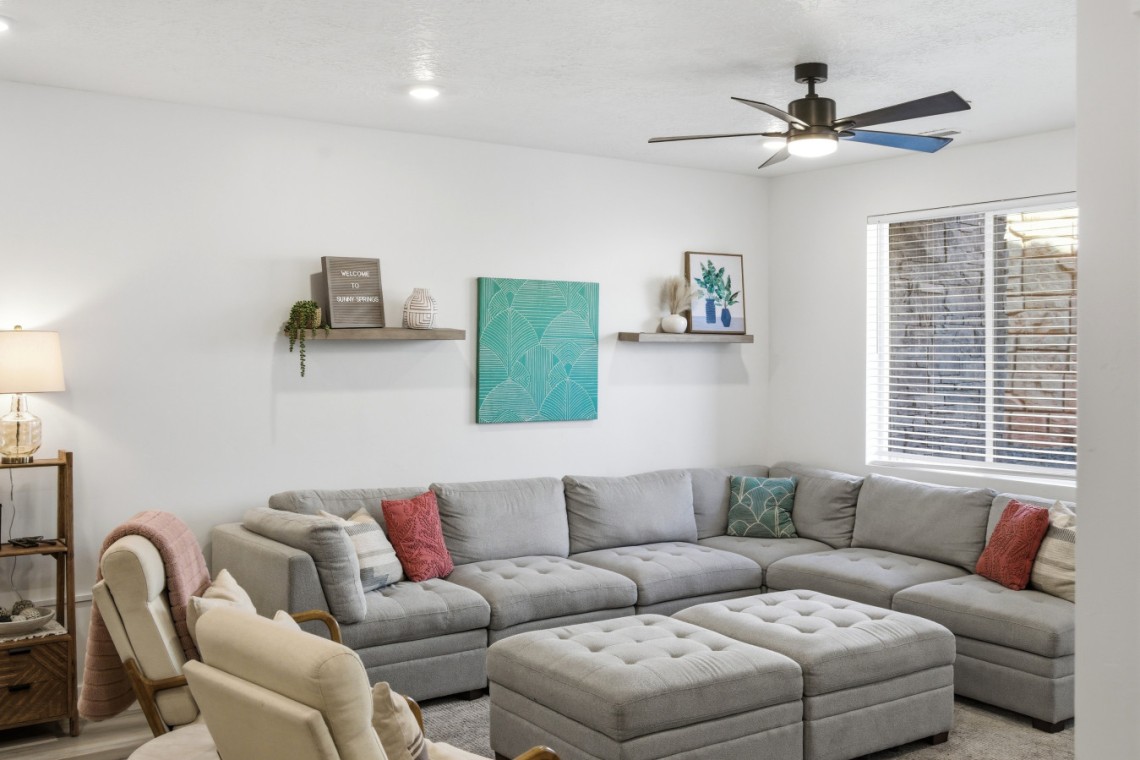 Bright living room with a gray sectional sofa, tufted ottoman, and decorative teal art on the wall above two floating shelves.