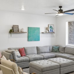 Bright living room with a gray sectional sofa, tufted ottoman, and decorative teal art on the wall above two floating shelves.