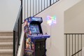 NFL Blitz arcade cabinet beside a neon 'GAME ROOM' sign in a stairwell, with metal railing and carpeted stairs nearby.