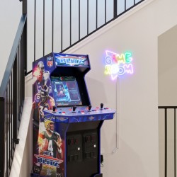 NFL Blitz arcade cabinet beside a neon 'GAME ROOM' sign in a stairwell, with metal railing and carpeted stairs nearby.