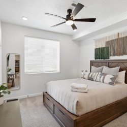 Wood bed with beige bedding and patterned throw pillows in a bright modern bedroom, ceiling fan above.
