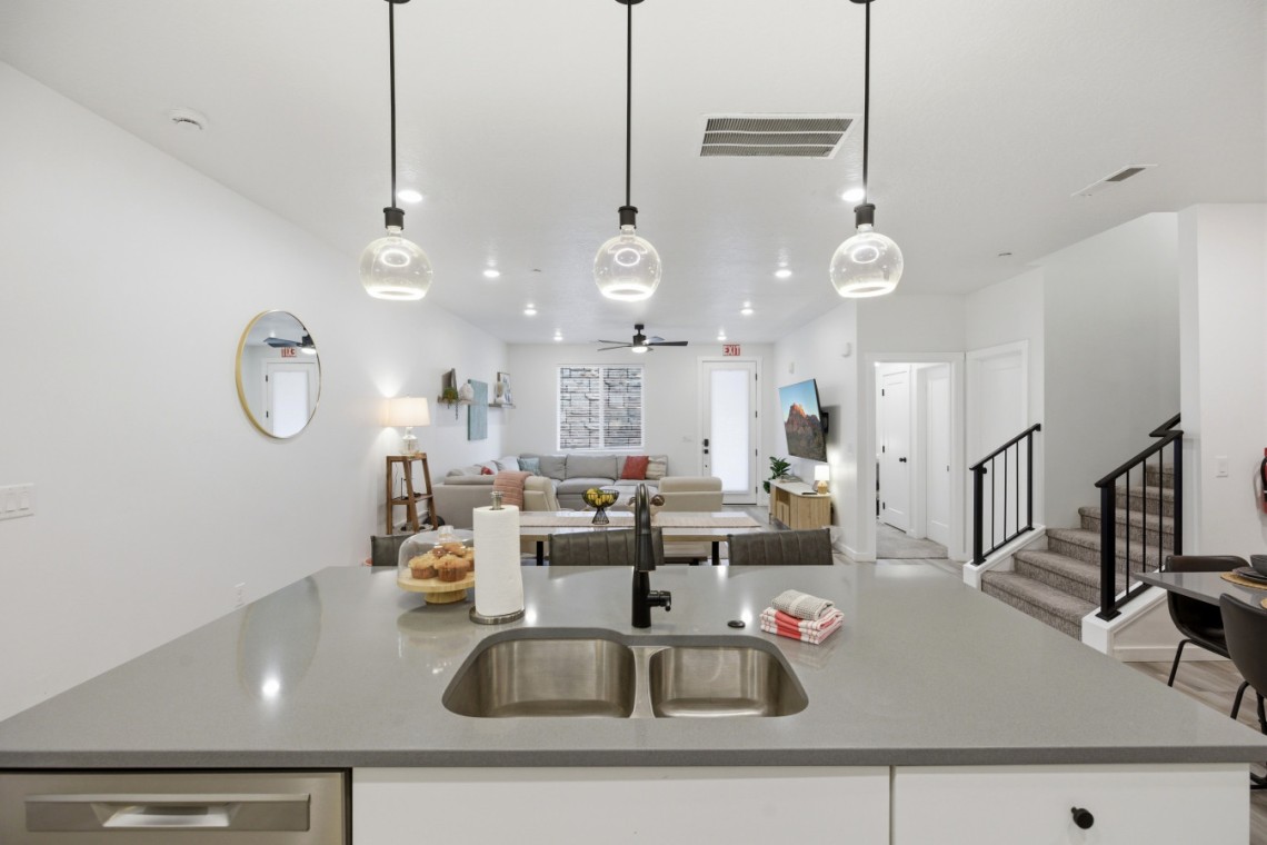 Open-concept kitchen and living area with a gray island, double sink, and pendant globe lights overlooking a cozy seating space.