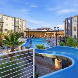 Outdoor pool area at a modern apartment complex, featuring a bright blue winding water slide, lounge chairs, and tropical landscaping under a clear sky.