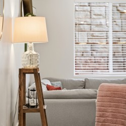 Living room scene with a glass-lit lamp on a wooden ladder-style side table beside a gray sofa and pink throw blanket on the armrest.
