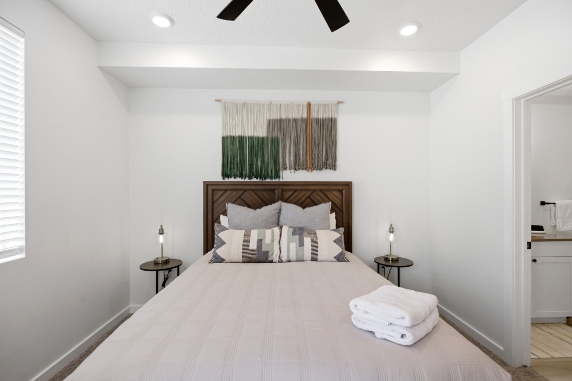 Queen bed with a wooden headboard, neutral quilt, patterned throw pillows, and folded towels on the foot of the bed; twin side tables with lamps flank the bed; decorative wall textile hangs above the headboard.