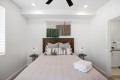 Queen bed with a wooden headboard, neutral quilt, patterned throw pillows, and folded towels on the foot of the bed; twin side tables with lamps flank the bed; decorative wall textile hangs above the headboard.
