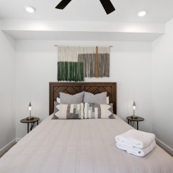 Queen bed with a wooden headboard, neutral quilt, patterned throw pillows, and folded towels on the foot of the bed; twin side tables with lamps flank the bed; decorative wall textile hangs above the headboard.