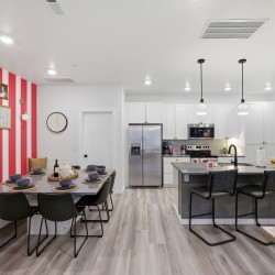 Open-concept kitchen and dining area with a pink striped accent wall, framed art, and a long dining table set for eight beside a white-cabinet kitchen and island with bar stools.