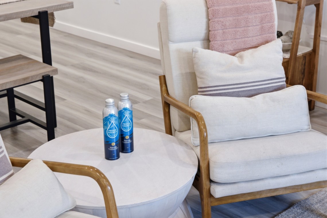 Two blue bottled waters sit on a round white side table beside a cream upholstered armchair with wooden arms in a cozy living room setting.
