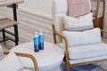 Two blue bottled waters sit on a round white side table beside a cream upholstered armchair with wooden arms in a cozy living room setting.