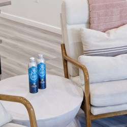 Two blue bottled waters sit on a round white side table beside a cream upholstered armchair with wooden arms in a cozy living room setting.