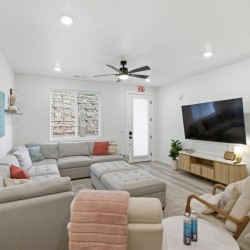 Bright living room with a large light-gray sectional sofa, matching ottoman, and pastel throw pillows facing a wall-mounted flat-screen TV.