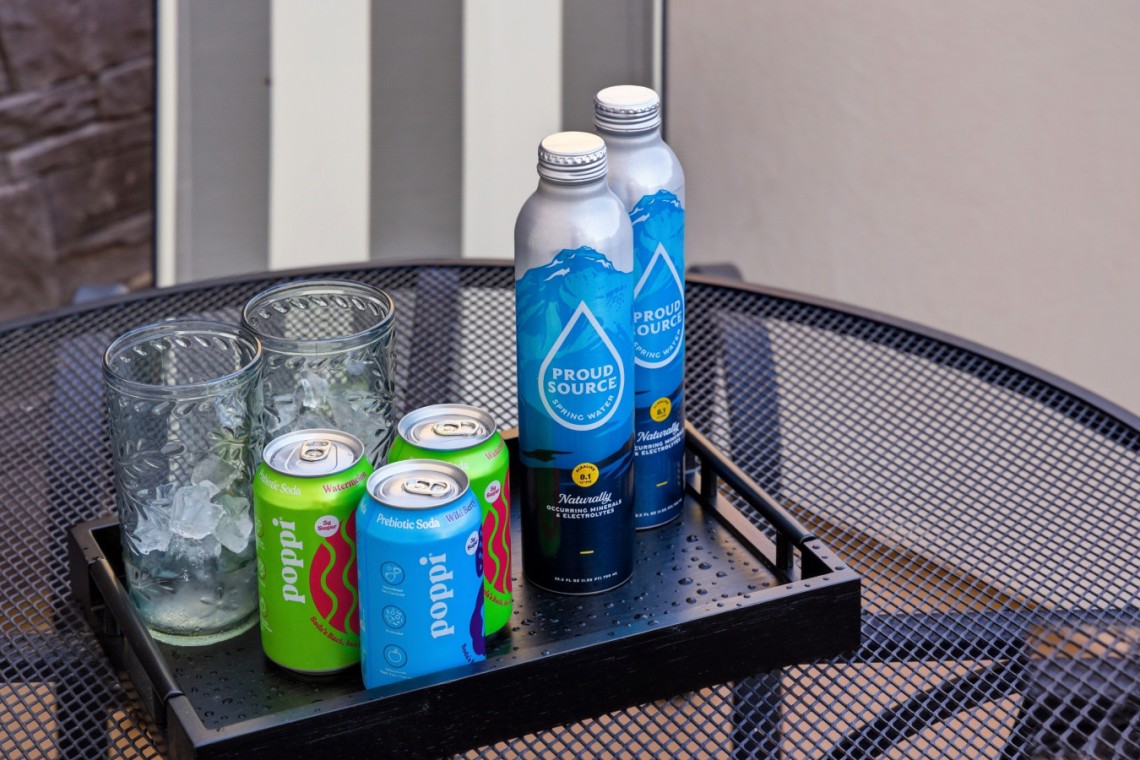 Tray with ice-filled glasses, two green and blue Poppi soda cans, and two Proud Source water bottles on a perforated metal table outside.