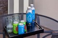Tray with ice-filled glasses, two green and blue Poppi soda cans, and two Proud Source water bottles on a perforated metal table outside.