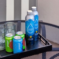 Tray with ice-filled glasses, two green and blue Poppi soda cans, and two Proud Source water bottles on a perforated metal table outside.