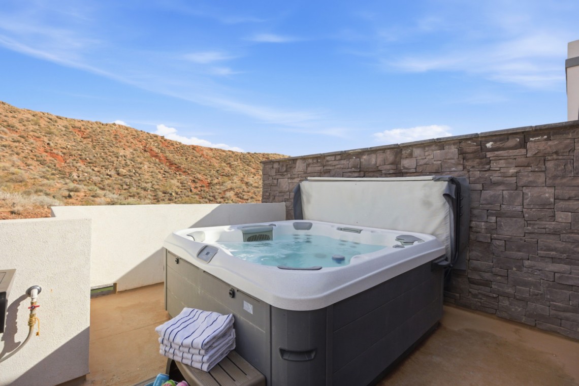 Outdoor hot tub on a concrete patio beside a stone privacy wall, with desert hills and a blue sky beyond.