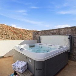 Outdoor hot tub on a concrete patio beside a stone privacy wall, with desert hills and a blue sky beyond.