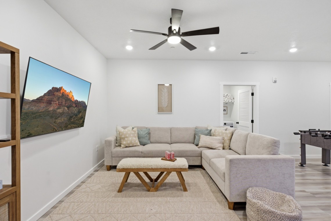 L-shaped beige sofa with decorative pillows facing a wall-mounted flat-screen TV showing a canyon landscape, in a bright living room.