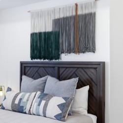Bedroom with a dark wood headboard, layered pillows, and a large gradient textile wall hanging above the bed.