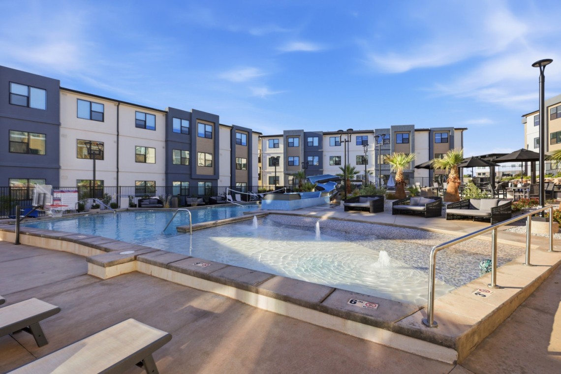 Outdoor pool area of a modern apartment complex with lounge chairs, palm trees, and shaded seating under umbrellas.