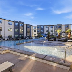 Outdoor pool area of a modern apartment complex with lounge chairs, palm trees, and shaded seating under umbrellas.