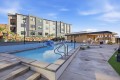 Outdoor swimming pool in a modern apartment complex, with lounge chairs, a low clubhouse, and a blue sky overhead