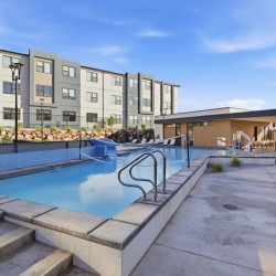 Outdoor swimming pool in a modern apartment complex, with lounge chairs, a low clubhouse, and a blue sky overhead