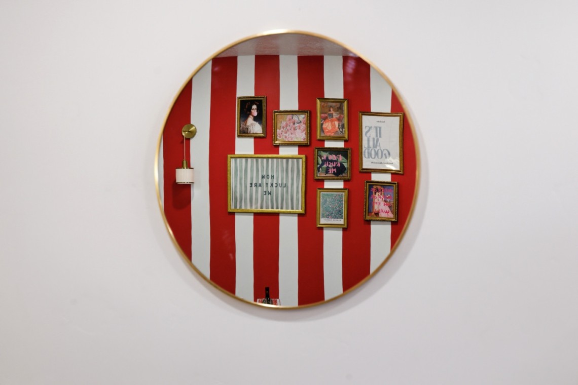 Round wall mirror reflecting a red-and-white striped gallery wall with multiple framed pictures and a small wall lamp on the left.