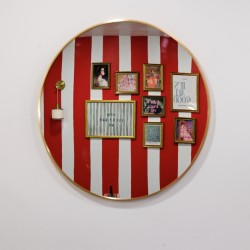 Round wall mirror reflecting a red-and-white striped gallery wall with multiple framed pictures and a small wall lamp on the left.