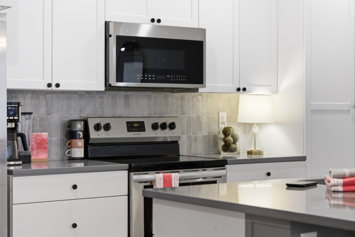 Modern kitchen with stainless steel microwave above a range, white cabinets, and a gray tiled backsplash.