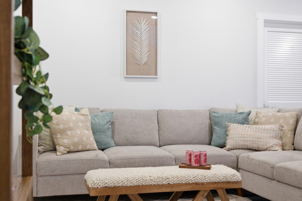 Beige sectional sofa with multiple decorative pillows in a bright living room; a wooden coffee table with a tray of pink drinks sits in front.