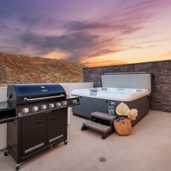 Patio at sunset with a black gas grill and a large hot tub against a stone wall.