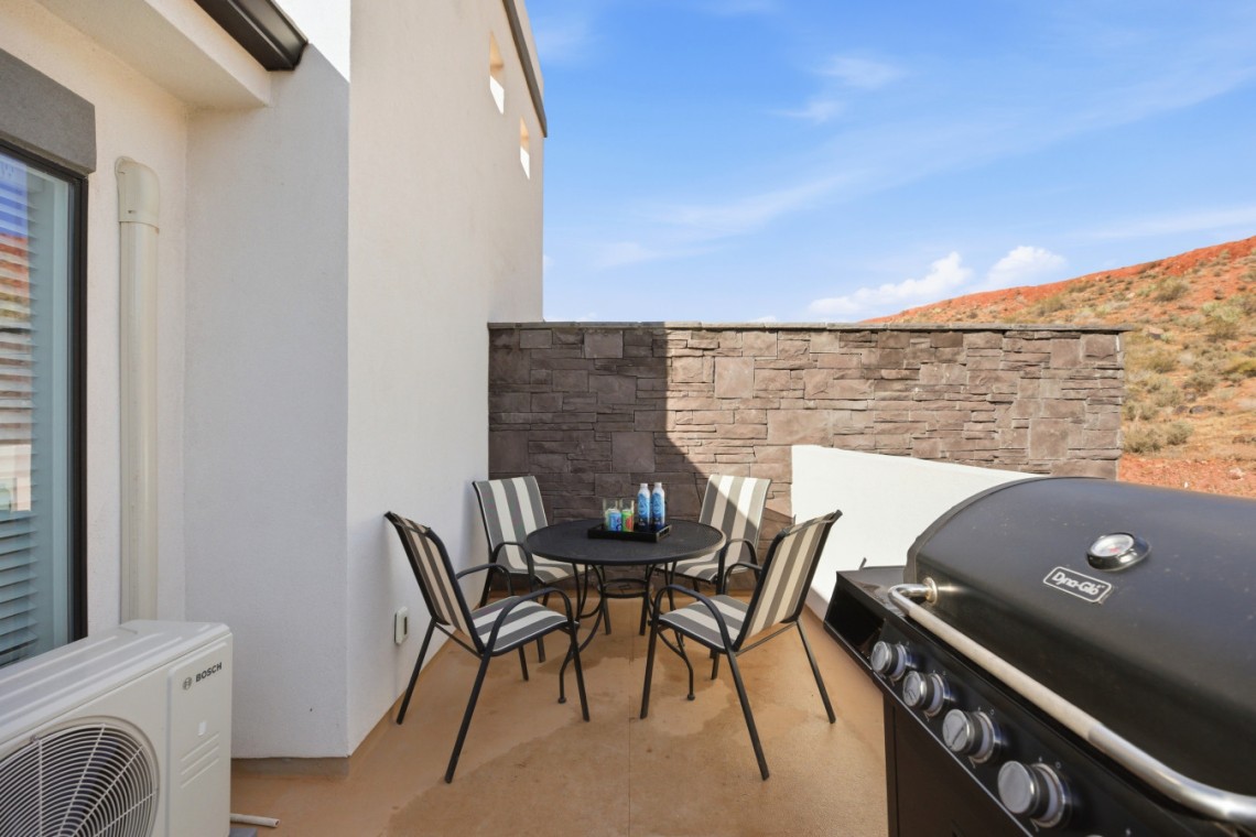 Outdoor balcony seating area with a round table and six striped chairs, a grill on the right, and a stone wall backdrop.