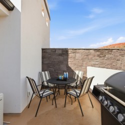 Outdoor balcony seating area with a round table and six striped chairs, a grill on the right, and a stone wall backdrop.