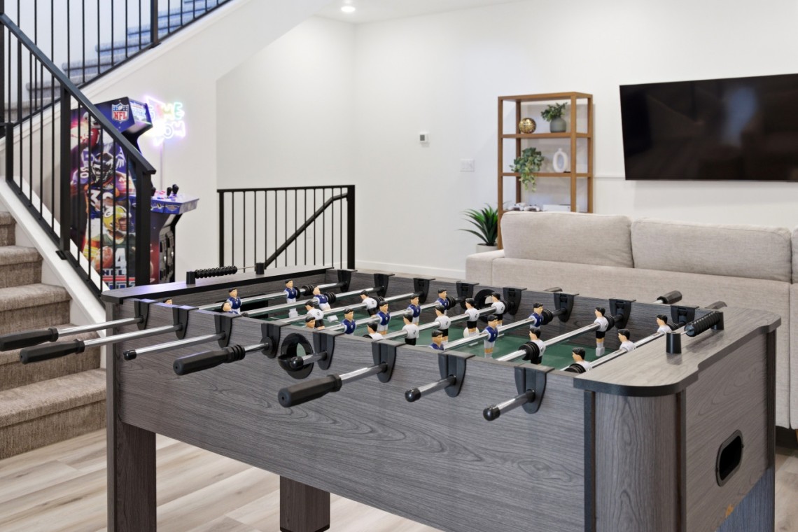 Foosball table in a modern basement recreation room with stairs, arcade machine, and a beige sofa with a wall-mounted TV in the background