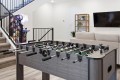 Foosball table in a modern basement recreation room with stairs, arcade machine, and a beige sofa with a wall-mounted TV in the background